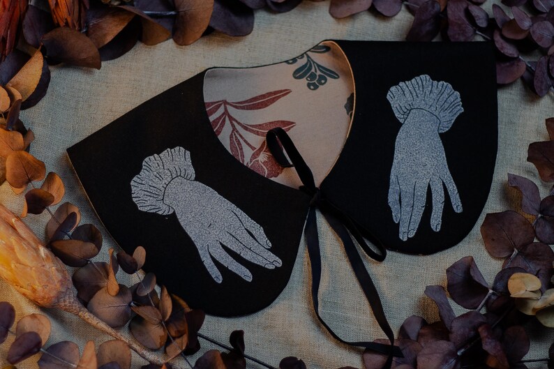 May include: A black collar with a floral patterned lining and a black ribbon tie. The collar features a silver-colored hand design on each side. The background includes dried leaves and a neutral-colored fabric.