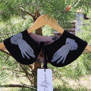 May include: A black detachable collar with a silver hand design, hanging on a wooden hanger. The collar has a floral patterned lining and a black ribbon tie. A white tag with a botanical illustration hangs below.