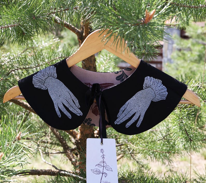 May include: A black detachable collar with a silver hand design, hanging on a wooden hanger. The collar has a floral patterned lining and a black ribbon tie. A white tag with a botanical illustration hangs below.