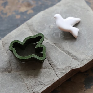 May include: A dark green bird-shaped cookie cutter with a layered texture and a smooth, glossy white ceramic bird figurine. The cookie cutter has an open center and defined edges, while the white bird is solid with a curved wing and tail.
