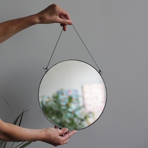 Puede incluir: Un espejo redondo con marco negro y cuerda para colgar. El espejo refleja una imagen borrosa de plantas verdes.