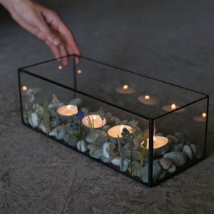 May include: A rectangular glass terrarium with a black metal frame, filled with smooth grey stones, dried flowers, and lit tea light candles. The candles cast a warm glow, creating a serene and decorative display.