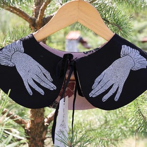 May include: Black collar with a hand design in silver glitter, hanging on a wooden hanger. The collar has a black tie closure and a patterned lining. The background is blurred with green foliage.