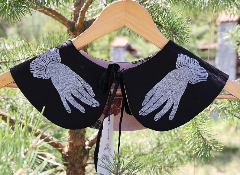 May include: Black collar with a hand design in silver glitter, hanging on a wooden hanger. The collar has a black tie closure and a patterned lining. The background is blurred with green foliage.