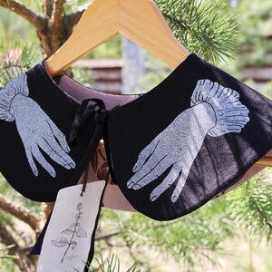 May include: Black collar with a hand design in white. The collar is hanging on a wooden hanger. The collar has a pink lining and a black tie. A tag with a botanical illustration hangs from the collar.