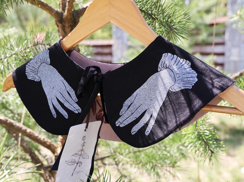 May include: Black collar with a hand design in white. The collar is hanging on a wooden hanger. The collar has a pink lining and a black tie. A tag with a botanical illustration hangs from the collar.