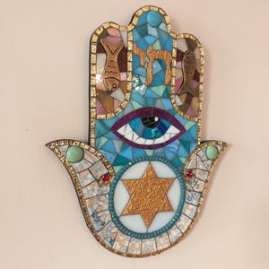 Puede incluir: Un colgante de pared Hamsa con un diseño de mosaico en tonos azul, blanco y dorado. La pieza incluye una Estrella de David central, un ojo estilizado, dos figuras de peces y la letra hebrea Chai, bordeada por un marco metálico dorado.
