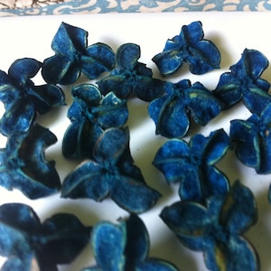 May include: A collection of small, blue, flower-shaped objects with a textured appearance. The objects are scattered across a white surface, with some showing hints of yellow. The overall aesthetic suggests a craft or decorative item.