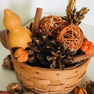 May include: A round woven wooden basket filled with a seasonal arrangement of a smooth yellow gourd, brown pinecones, orange rattan spheres, cinnamon sticks, and dried botanical elements. The basket features a light brown finish and horizontal slats.