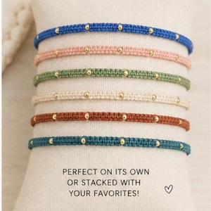 May include: Six woven cord bracelets in blue, pink, green, cream, rust, and teal, each featuring small gold-toned beads spaced along the band. The text "PERFECT ON ITS OWN OR STACKED WITH YOUR FAVORITES!" and a small heart icon are printed below.