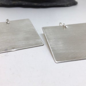 以下が含まれることがあります： ブラッシュ仕上げのシルバーの正方形のイヤリングのペア。イヤリングはシンプルでエレガントです。