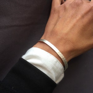 May include: A silver bracelet with a hammered texture. The bracelet is worn on a person's wrist.