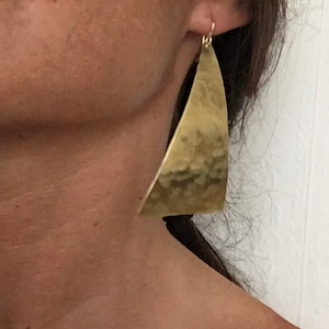 May include: A pair of large, gold-coloured earrings with a hammered texture. The earrings have a triangular shape with a curved edge, hanging from a simple hook. The earrings are a statement piece, adding a touch of modern elegance.