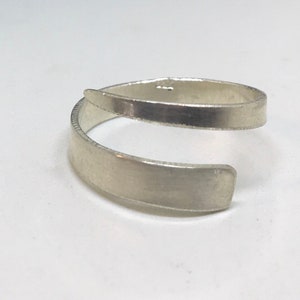May include: A silver ring with a unique design, featuring a band that wraps around the finger twice. The ring is made of smooth, polished metal and has a simple, modern aesthetic.