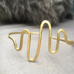 Op de afbeelding: Een goudkleurige manchetarmband met een golvend, abstract ontwerp. De armband heeft een doorlopende lijn die buigt en lussen maakt, waardoor een unieke en moderne esthetiek ontstaat. Het metaal heeft een matte afwerking.