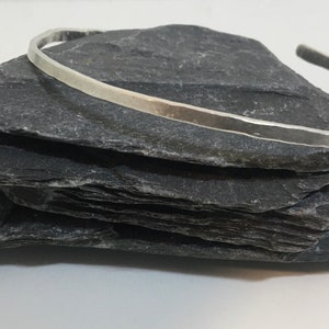 May include: A silver bangle bracelet with a hammered finish. The bracelet is displayed on a stack of dark grey slate tiles.