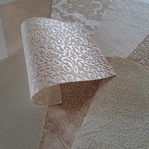 May include: A selection of wallpaper samples in neutral shades. The samples feature textured and patterned designs, predominantly in beige and cream tones. One sample is rolled, revealing an embossed, swirling pattern.