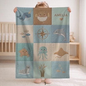 Può includere: Una coperta per bambini con un design patchwork con creature marine come un narvalo, un delfino e una medusa. La coperta è nei toni del blu, beige e marrone chiaro, con il nome "AMELIA" stampato.