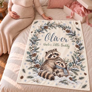 May include: A cream-colored fleece blanket featuring a printed illustration of two raccoons nestled in snow, surrounded by a wreath of pine branches, pinecones, and snowflakes. The text reads Oliver and Uncle's Little Buddy.