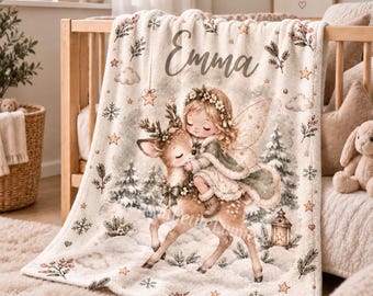 May include: A cream-colored fleece blanket featuring the name Emma in cursive script. The design shows a fairy with wings hugging a fawn in a snowy forest setting with pine trees, snowflakes, stars, and a lantern. The print includes soft green and brown tones.