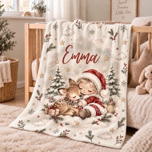 Könnte beinhalten: Eine cremefarbene Fleecedecke mit einer weihnachtlichen Illustration eines Kindes mit roter Weihnachtsmütze, das ein kleines Reh umarmt. Das Design zeigt Tannenbäume, Schneeflocken, Sterne, Beeren und eine Laterne. Der Name Emma ist in Rot aufgedruckt.