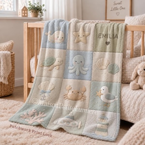 Könnte beinhalten: Eine weiche, flauschige Babydecke mit einem Raster aus gestickten nautischen und maritimen Motiven, darunter ein Wal, ein Seestern, eine Schildkröte, ein Oktopus, Muscheln, eine Krabbe, eine Möwe, Korallen, ein U-Boot und ein Leuchtturm, mit dem Namen EMILIA und einem Herz.