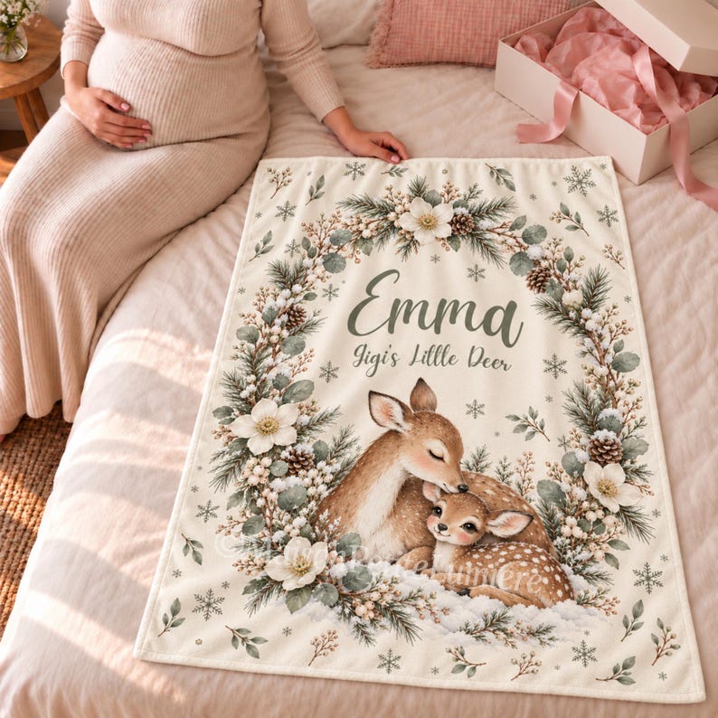 May include: A cream-colored rectangular blanket featuring a printed illustration of two deer nestled within a wreath of pine branches, white flowers, and pinecones. The text reads Emma and Jigi's Little Deer, surrounded by small snowflakes.