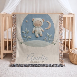 Pode incluir: Um cobertor de bebê bege com um design circular azul com um ursinho de pelúcia em um macacão com capuz, uma lua crescente e uma estrela. O cobertor tem uma borda com franjas e o nome "Amelia" e a data "01.01.2025" são bordados.