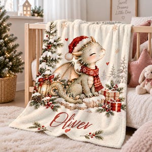 Könnte beinhalten: Eine cremefarbene Fleecedecke mit einer aufgedruckten Illustration eines kleinen Drachen, der eine rote Weihnachtsmütze und einen roten gemusterten Schal trägt. Das Design zeigt einen verschneiten Weihnachtsbaum, eine Laterne, Geschenke und den Namen Oliver.