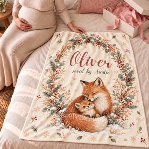 May include: A cream-colored fleece blanket featuring a printed wreath of pine branches, red berries, and snowflakes. The text reads Oliver and Loved by Auntie. A large adult fox and a small fox kit are nestled together at the bottom center.