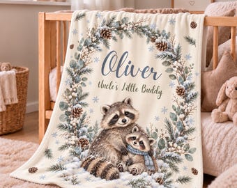 May include: A cream-colored fleece blanket featuring a printed design of two raccoons surrounded by a winter wreath of pine branches, pinecones, and berries. The text reads Oliver and Uncle's Little Buddy in dark blue and green script.