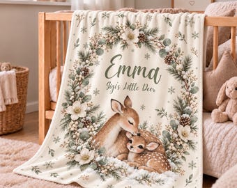 May include: A cream-coloured fleece blanket featuring a printed wreath of pine branches, white flowers, and snowflakes. The centre displays the name Emma and the text Jigi's Little Deer above an illustration of two deer resting together.