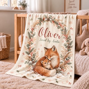 Personalized fox baby blanket with name held up in a warm nursery, mama fox curled around her smiling kit inside a snowy red berry and pine wreath