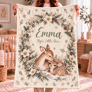 May include: A cream-colored fleece blanket featuring a central illustration of a mother deer and fawn nestled in snow. A wreath of pine branches, white flowers, and pinecones surrounds the text Emma and Jigi's Little Deer.