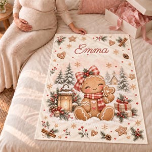 Gingerbread baby blanket with name held up in a nursery, showing a gingerbread girl with a red gingham bow and scarf among snowy pine trees, a lantern and presents.