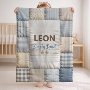 May include: A patchwork baby blanket in shades of blue, beige, and white. The blanket features the name "LEON" and the phrase "Simply Loved" with two star graphics. The blanket is held up by a person.
