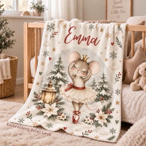 May include: A cream-colored fleece blanket featuring the name Emma in red script. The design includes a mouse wearing a tutu and crown, surrounded by winter elements like pine trees, snowflakes, red berries, white poinsettias, and a lantern.