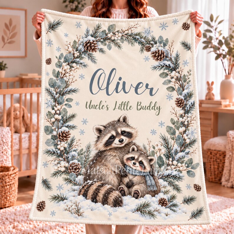 May include: A cream-colored fleece blanket featuring a printed winter wreath of pine branches, pinecones, and white berries. The text reads Oliver and Uncle's Little Buddy above an illustration of two raccoons in the snow.