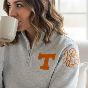 May include: A light gray quarter-zip pullover sweatshirt featuring an orange letter T on the left chest and an orange embroidered monogram on the left shoulder. The garment has long sleeves, a high collar with a silver zipper, and a soft texture.