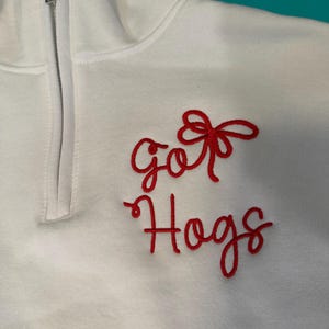 May include: A white quarter-zip pullover featuring red embroidered text that reads "go" above a red bow and the word "Hogs" written in a cursive script below. The garment has a high collar with a silver-toned metal zipper closure at the neck.