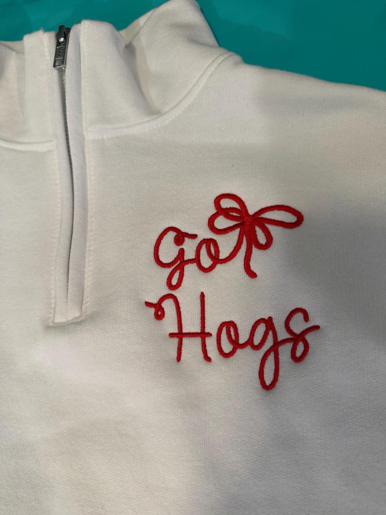 May include: A white quarter-zip pullover featuring red embroidered text that reads "go" above a red bow and the word "Hogs" written in a cursive script below. The garment has a high collar with a silver-toned metal zipper closure at the neck.