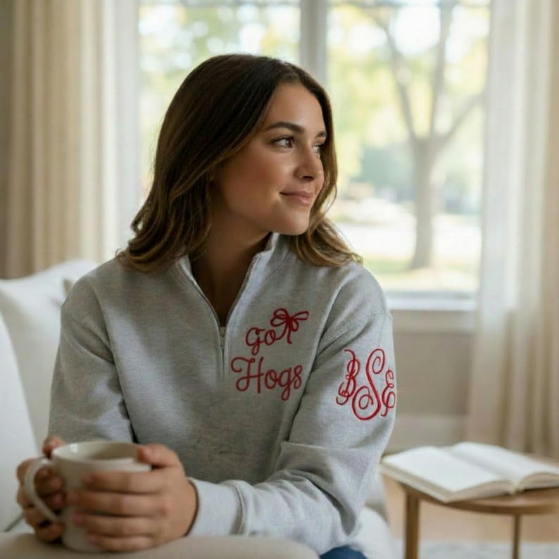 May include: A light gray quarter-zip sweatshirt featuring red embroidered text that reads Go Hogs with a bow graphic above the text and a red embroidered monogram on the sleeve. The garment has a high collar and long sleeves.