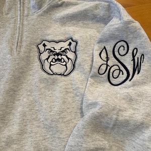 May include: A light gray quarter-zip sweatshirt with a bulldog logo patch on the chest and embroidered initials on the sleeve. The bulldog patch is in shades of gray and navy blue.