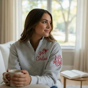 May include: A light gray quarter-zip sweatshirt with the words "Go Cardinals" embroidered in red, along with a red bow and monogram. The sweatshirt has a high collar and a zipper. The person is holding a white mug.