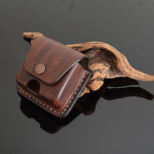May include: A brown leather case with a flap closure and a small metal button. The case is stitched with white thread and has a worn, vintage look.