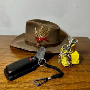 Puede incluir: Un sombrero de vaquero marr&oacute;n con una pluma, un llavero de cuero negro con una llave de coche y una figura de metal amarilla de un vaquero montando a caballo.