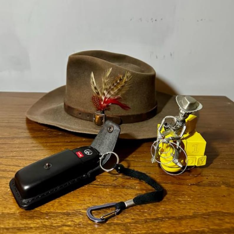 Puede incluir: Un sombrero de vaquero marr&oacute;n con una pluma, un llavero de cuero negro con una llave de coche y una figura de metal amarilla de un vaquero montando a caballo.