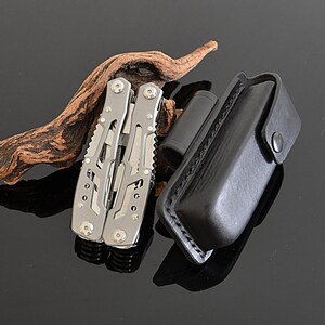 Peut inclure: Un multi-outil argent&eacute; avec plusieurs fonctions, &agrave; c&ocirc;t&eacute; d'un &eacute;tui en cuir noir. L'outil est ouvert, r&eacute;v&eacute;lant divers instruments. Un objet cylindrique noir et un morceau de bois flott&eacute; sont &eacute;galement pr&eacute;sents.