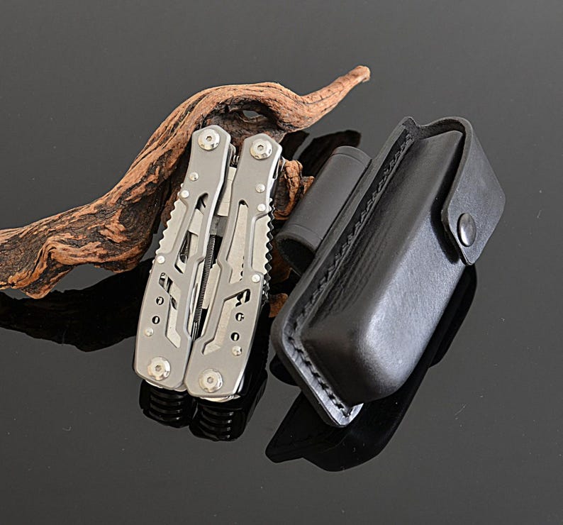 Peut inclure: Un multi-outil argent&eacute; avec plusieurs fonctions, &agrave; c&ocirc;t&eacute; d'un &eacute;tui en cuir noir. L'outil est ouvert, r&eacute;v&eacute;lant divers instruments. Un objet cylindrique noir et un morceau de bois flott&eacute; sont &eacute;galement pr&eacute;sents.