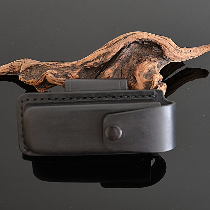 Peut inclure: Un &eacute;tui en cuir noir avec une fermeture &agrave; bouton-pression. L'&eacute;tui est con&ccedil;u pour contenir un petit outil ou un couteau. L'arri&egrave;re-plan pr&eacute;sente un morceau de bois flott&eacute;.